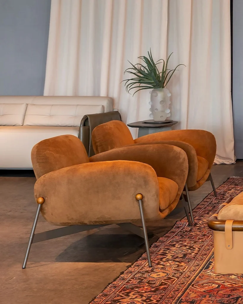 Luxo e Sofisticação: Elevato Casa chega ao site da Elevato 5 Poltrona Barista Salva com base metálica e assento estofado em tom caramelo, ambientada em sala moderna com tapete persa e sofá branco.