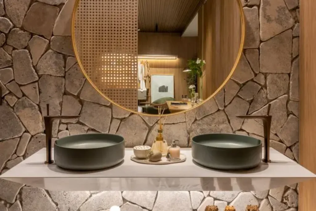 Cubas Deca: Design, Qualidade e Inovação para lavabos e banheiros 5 Banheiro elegante com parede de pedra natural, espelho redondo com moldura dourada, duas cubas deca apoio redondas verde torneiras altas em tom bronze sobre bancada branca.