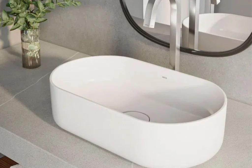 Cubas Deca: Design, Qualidade e Inovação para lavabos e banheiros 3 Cuba de apoio oval branca da Deca sobre bancada de pedra cinza, com torneira inox de parede, espelho com moldura preta e vaso com folhagens ao lado.