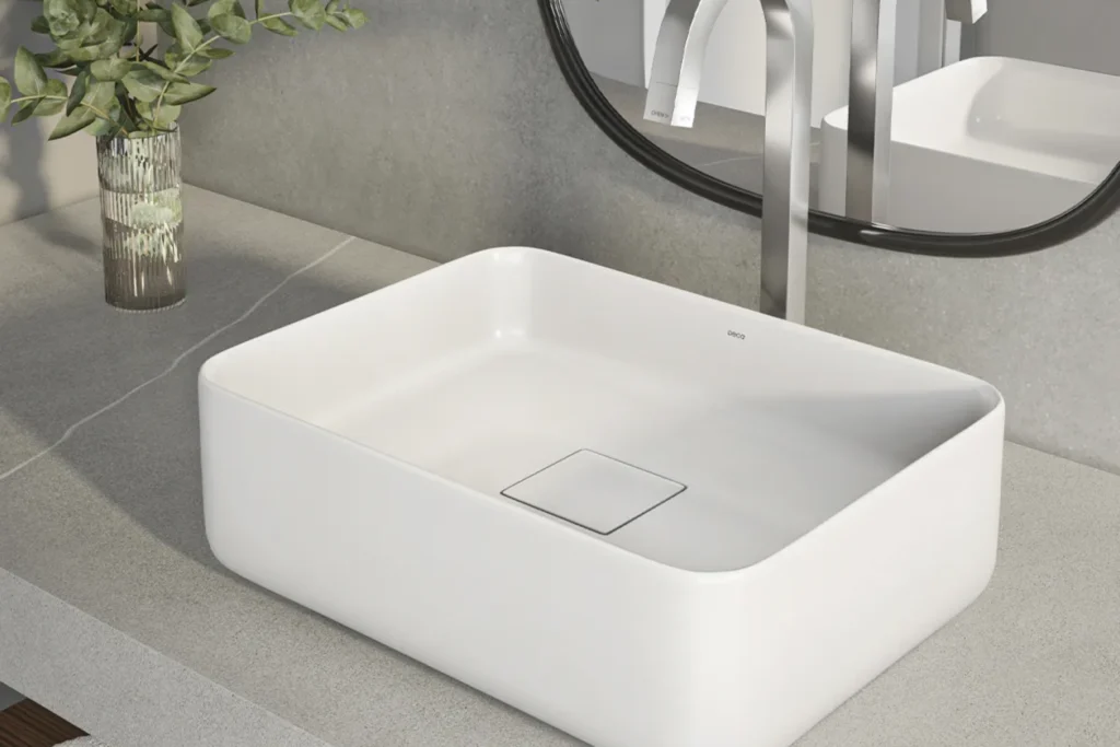 Cubas Deca: Design, Qualidade e Inovação para lavabos e banheiros 2 Cuba de apoio Deca Slim Quadrada branca com ralo oculto sobre bancada de pedra cinza, torneira inox de parede, espelho com moldura preta e vaso de vidro com folhagens ao lado.