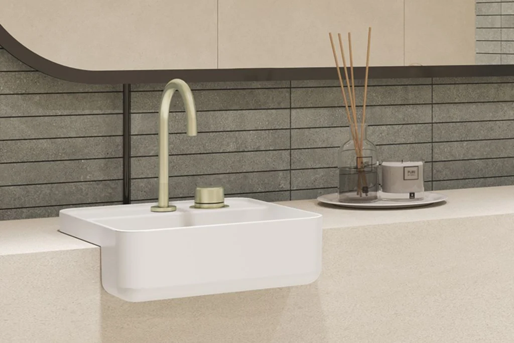 Cubas Deca: Design, Qualidade e Inovação para lavabos e banheiros 4 Banheiro moderno com cuba Deca de semiencaixe branco de cantos arredondados, torneira alta em tom champagne, bancada de pedra bege e fundo com revestimento cinza em filetes; ao lado, bandeja com difusor e vela.