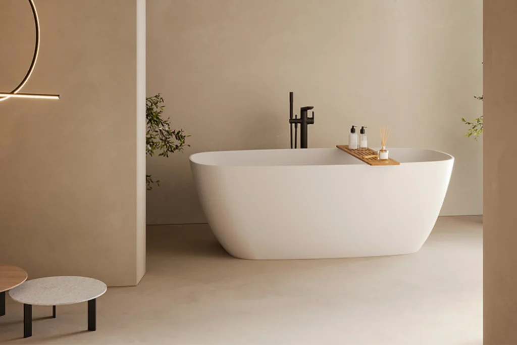 Banheira Sabbia: Conheça Modelos, Instalação e Inspirações 6 Banheira branca com bordas arredondadas, bandeja de madeira com frascos e difusor, misturador de piso preto ao fundo; ambiente minimalista em tons bege com mesas baixas e plantas discretas.