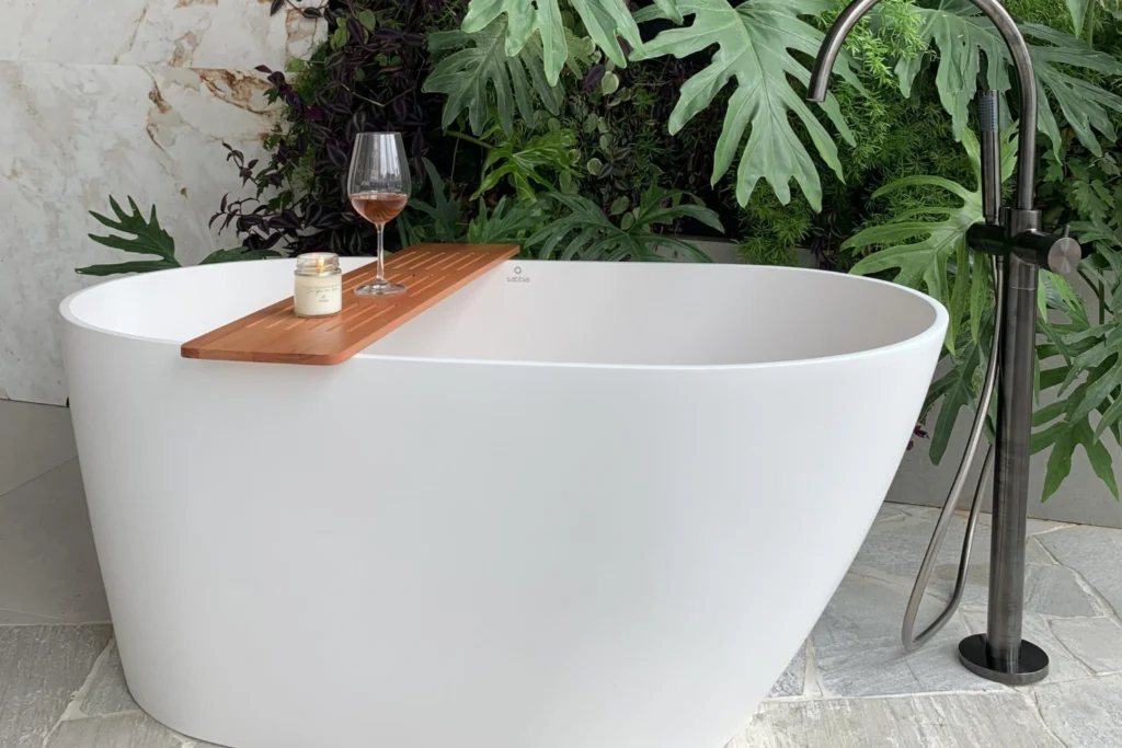 Banheira Sabbia: Conheça Modelos, Instalação e Inspirações 3 Banheira Sabbia Ilhéus branca, com acabamento fosco, bandeja de madeira apoiada com taça de vinho rosé e vela; ducha de piso preta ao lado, jardim vertical de folhagens e parede em mármore ao fundo.