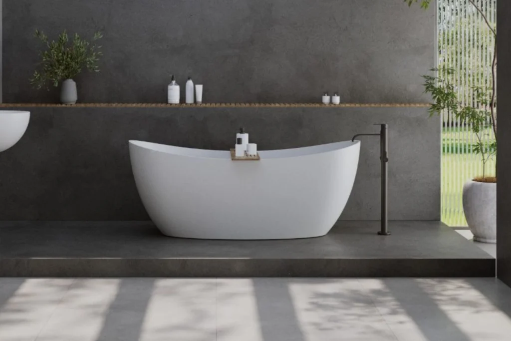 Banheira Sabbia: Conheça Modelos, Instalação e Inspirações 2 Banheiro moderno com banheira sabbia Sampa 170 moderno branca sobre base elevada em porcelanato escuro, prateleira de madeira com itens de banho, parede em cimento queimado, misturador de piso preto e jardim externo ao fundo.