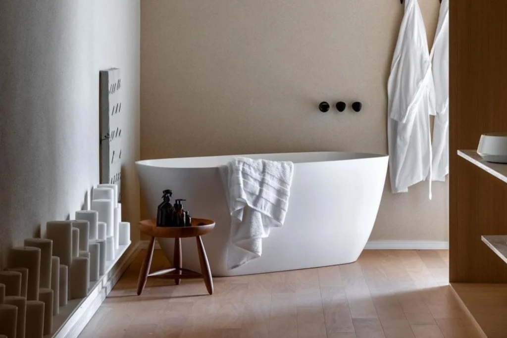 Banheira Sabbia: Conheça Modelos, Instalação e Inspirações 4 Banheira Sabbia Bahia de imersão freestanding branca, acabamento fosco, com toalha apoiada na borda; banquinho de madeira com frascos ao lado, parede bege com registros pretos, roupões pendurados e velas decorativas no rodapé.