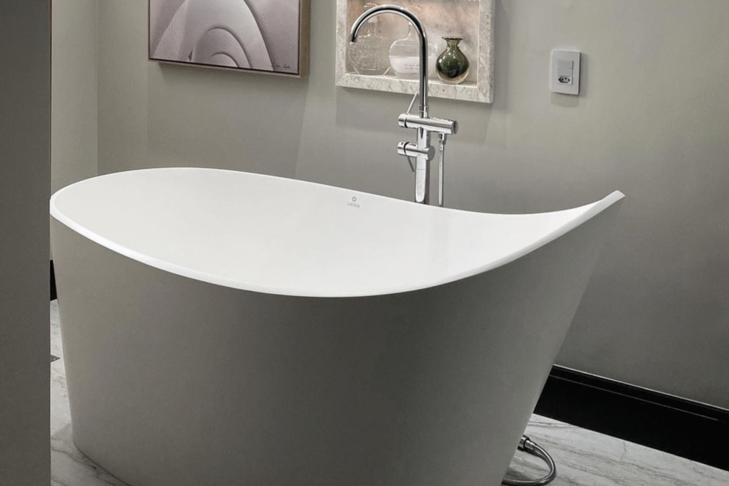 Banheira Sabbia: Conheça Modelos, Instalação e Inspirações 8 Banheira Sibba branca com borda elevada e assimétrica, instalada sobre piso marmorizado, com misturador de piso cromado e nicho decorativo na parede.