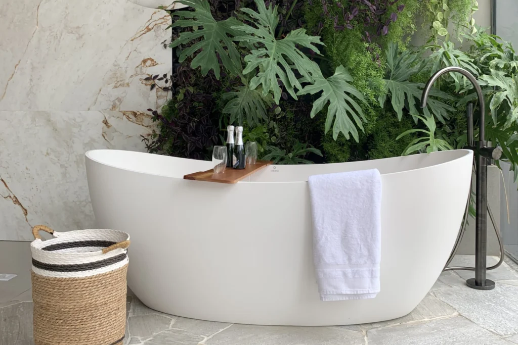 Banheira Sabbia: Conheça Modelos, Instalação e Inspirações 1 Imagem atual: Banheira Sabbia Sampa 170 branca com bandeja de madeira, taças e espumante, toalha branca apoiada na borda, ducha de piso ao lado, parede em mármore e jardim vertical com folhagens tropicais